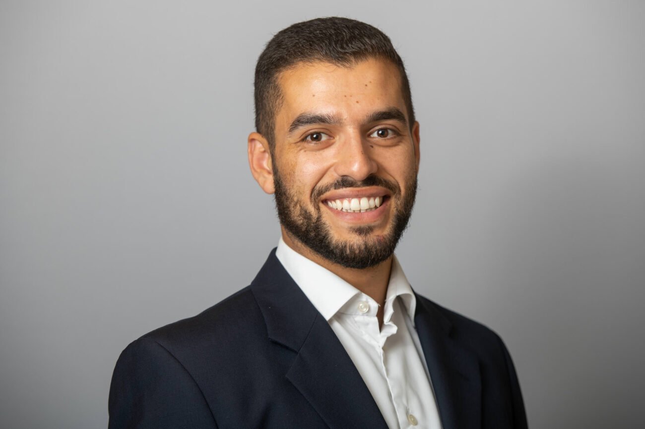 A smiling man with a beard in a dark suit and white shirt is standing against a plain gray background. James Gifford-Mead Photography - Event Photographer London