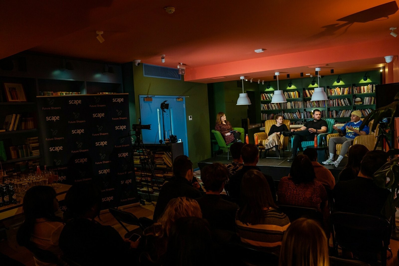 A panel of three people sitting in front of an audience at an indoor event. The background features a bookshelf with books and green walls. A person stands at a podium with a "scape" banner on the left. The lighting is dim and warm. James Gifford-Mead Photography - Event Photographer London