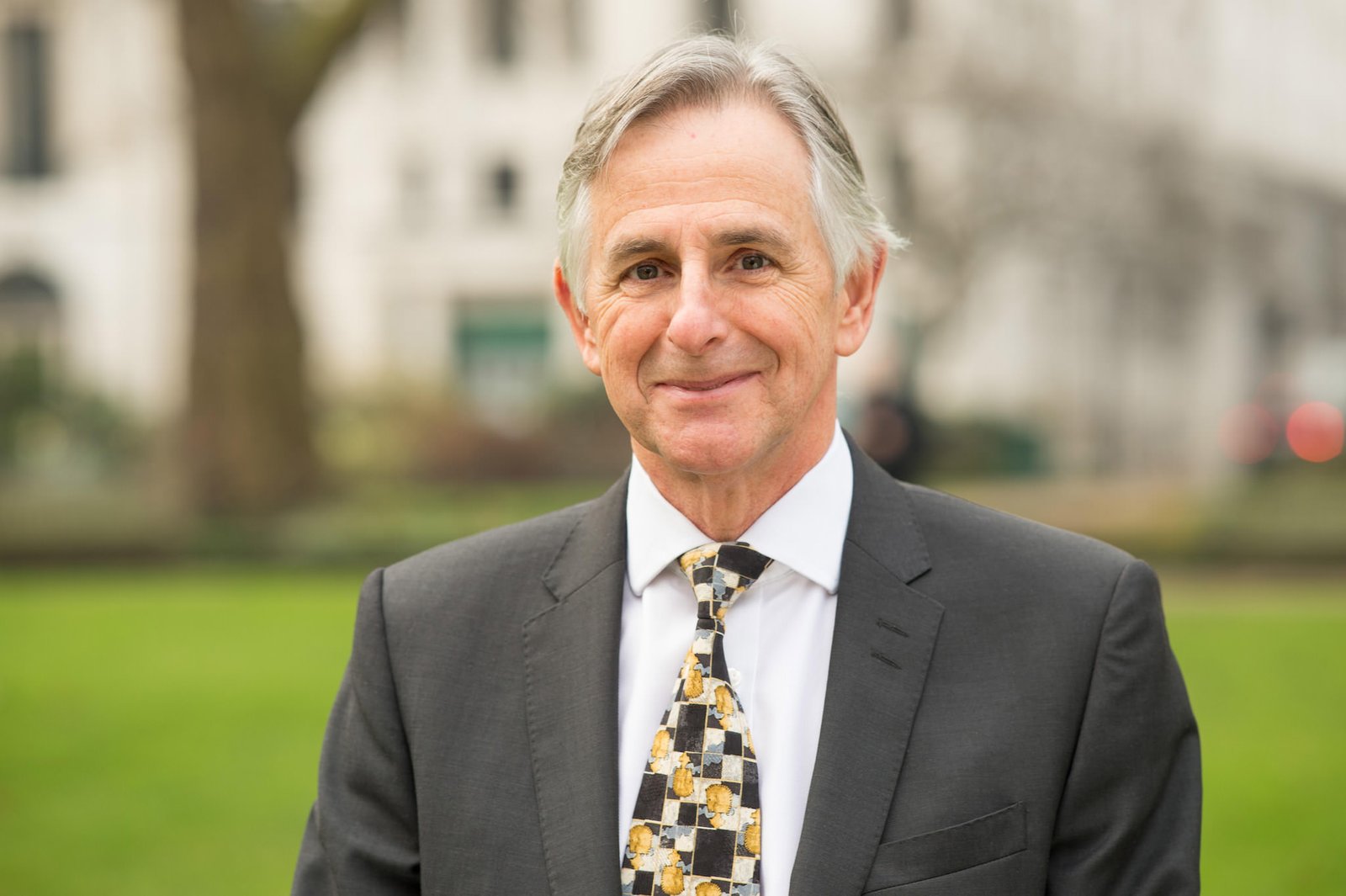 A man wearing a suit and patterned tie stands outdoors in a park. The background is blurred, featuring greenery and a tall building. James Gifford-Mead Photography - Event Photographer London