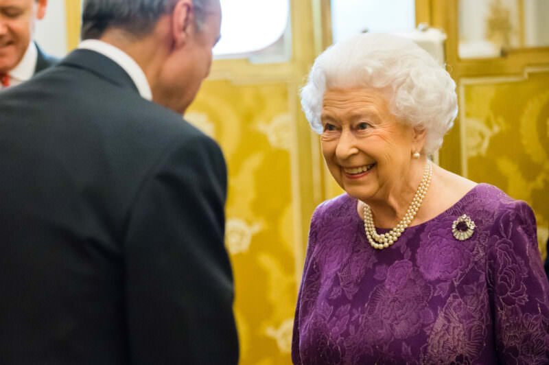 An older woman with white hair, wearing a purple dress and pearl necklace, smiles while speaking to a man in a suit. They are in an elegant room with golden decor