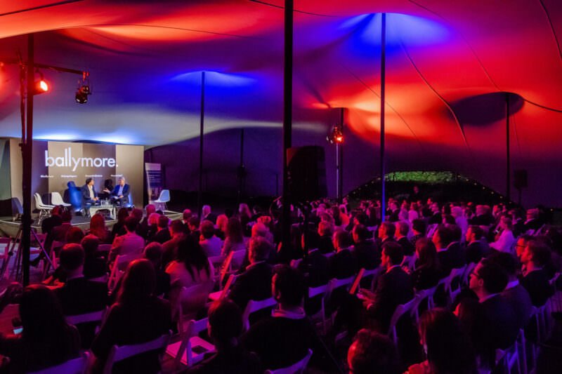 A well-attended indoor event with a large audience seated and listening to a group of speakers on stage. The ceiling is illuminated with vibrant red and blue lights, and a sign reading "ballymore" is visible behind the speakers