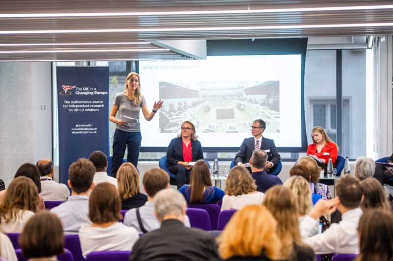 A woman speaks on stage in front of an audience, with three people seated beside her. A screen behind them displays a presentation titled "Where are the women?" and a banner on the left reads "The UK in a Changing Europe