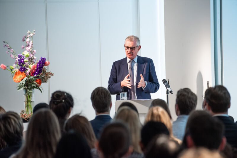 A man in a suit speaks passionately at a podium to a seated audience. A microphone is nearby, and a colorful bouquet of flowers decorates the space