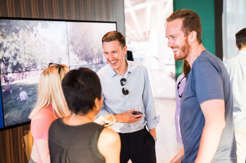 A group of five people stand together in a lively conversation. One man, holding a drink, is laughing. A large display screen is in the background, showing an outdoor scene. The atmosphere appears cheerful and social