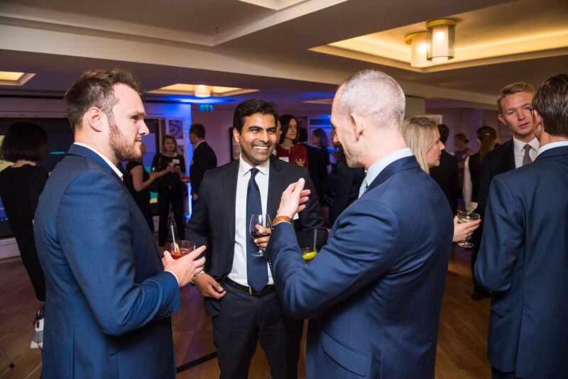 A group of people in formal attire are socializing at an indoor event. Three men in suits stand in the foreground, holding drinks and smiling. More attendees are visible in the background under warm lighting