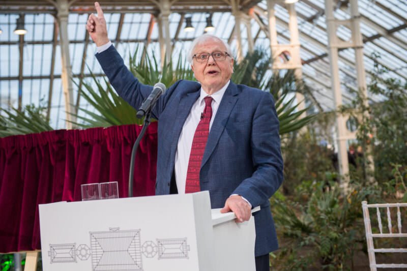 An elderly man in a suit and red tie stands at a podium, speaking animatedly with one arm raised. He is inside a greenhouse with large plants and glass panels visible in the background. A red curtain is partially visible to his left