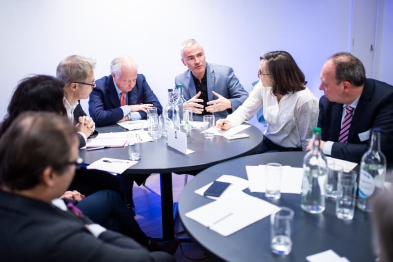 Seven people sit around a circular table in a discussion, with notebooks and bottles of water. A man gestures with his hands while speaking, and others listen attentively. The setting is a brightly lit conference room