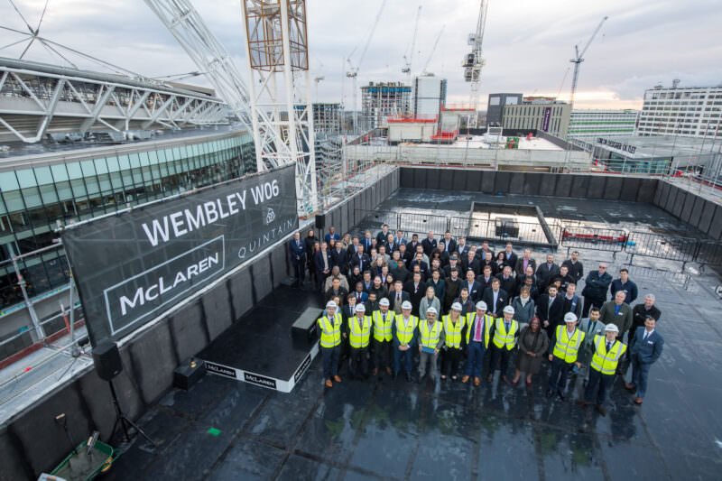 A large group of construction workers and professionals wearing safety vests and hard hats stand on a rooftop platform. Behind them, a tall crane and multiple buildings are visible. A banner reads "Wembley W06" with "McLaren" and "Quintain" logos