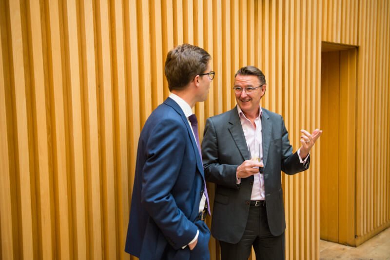 Two men in business attire stand talking in front of a wooden panel wall. The man on the left wears a blue suit and glasses, facing the man on the right, who wears a gray suit and gestures with his hand while holding a glass