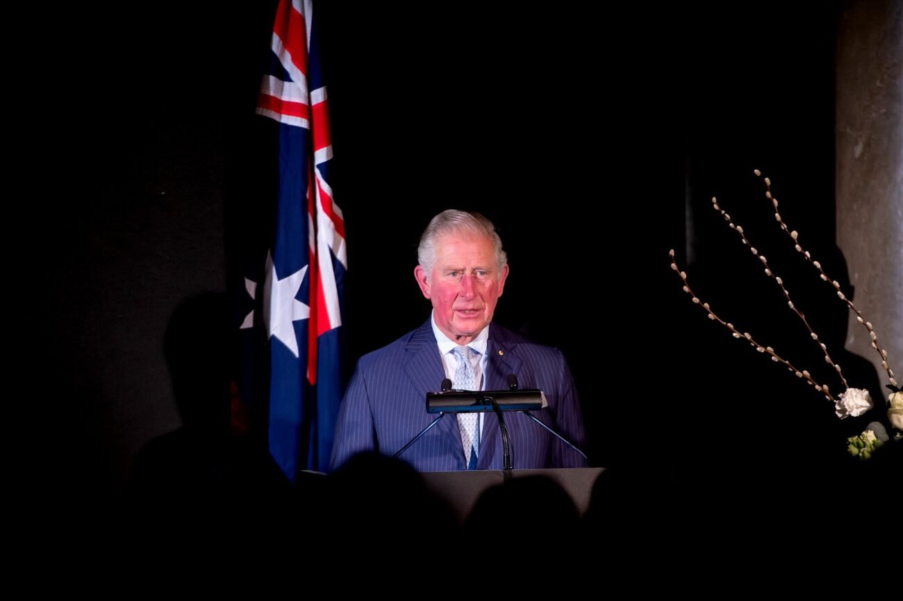 An older man stands at a podium, speaking into microphones. He wears a dark pinstripe suit and polka dot tie. An Australian flag is displayed behind him, and a branch with white flowers is visible to the right. The background is dark. James Gifford-Mead Photography - Event Photographer London