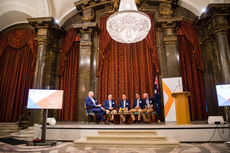 A panel of six people is seated on a stage in an ornate room with tall columns and a large chandelier. They are engaged in a discussion. A podium and a screen displaying graphics are on either side of the stage