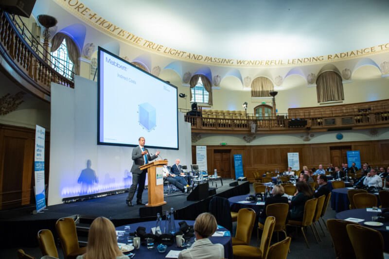 A man is presenting on stage in a large conference hall with a high ceiling and balcony. Attendees sit at tables with laptops and papers. A screen displays a slide titled "Notebook ODM." Busts and a quote adorn the walls