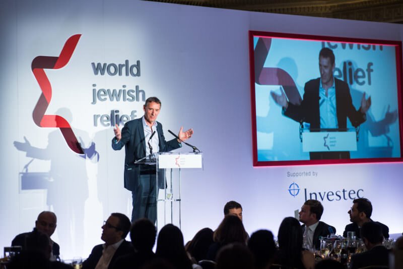 A speaker stands at a podium on a stage, addressing an audience at an event. The backdrop features the text "world jewish relief," and a screen displays the speaker. People are seated at tables in the foreground