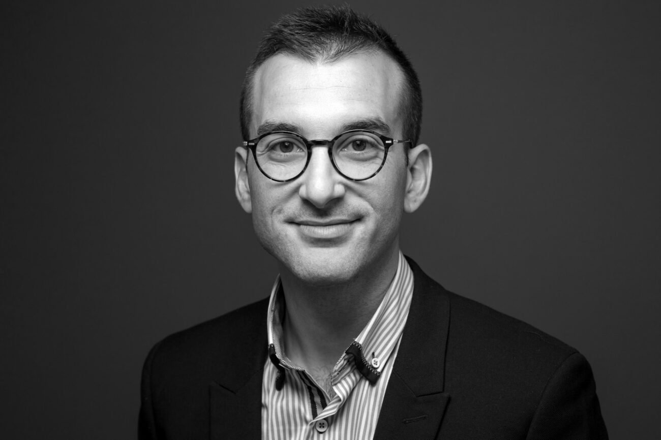 A black and white portrait of a person wearing glasses and a suit jacket over a striped shirt. The individual has short hair and is smiling slightly, set against a plain, dark background. James Gifford-Mead Photography - Event Photographer London