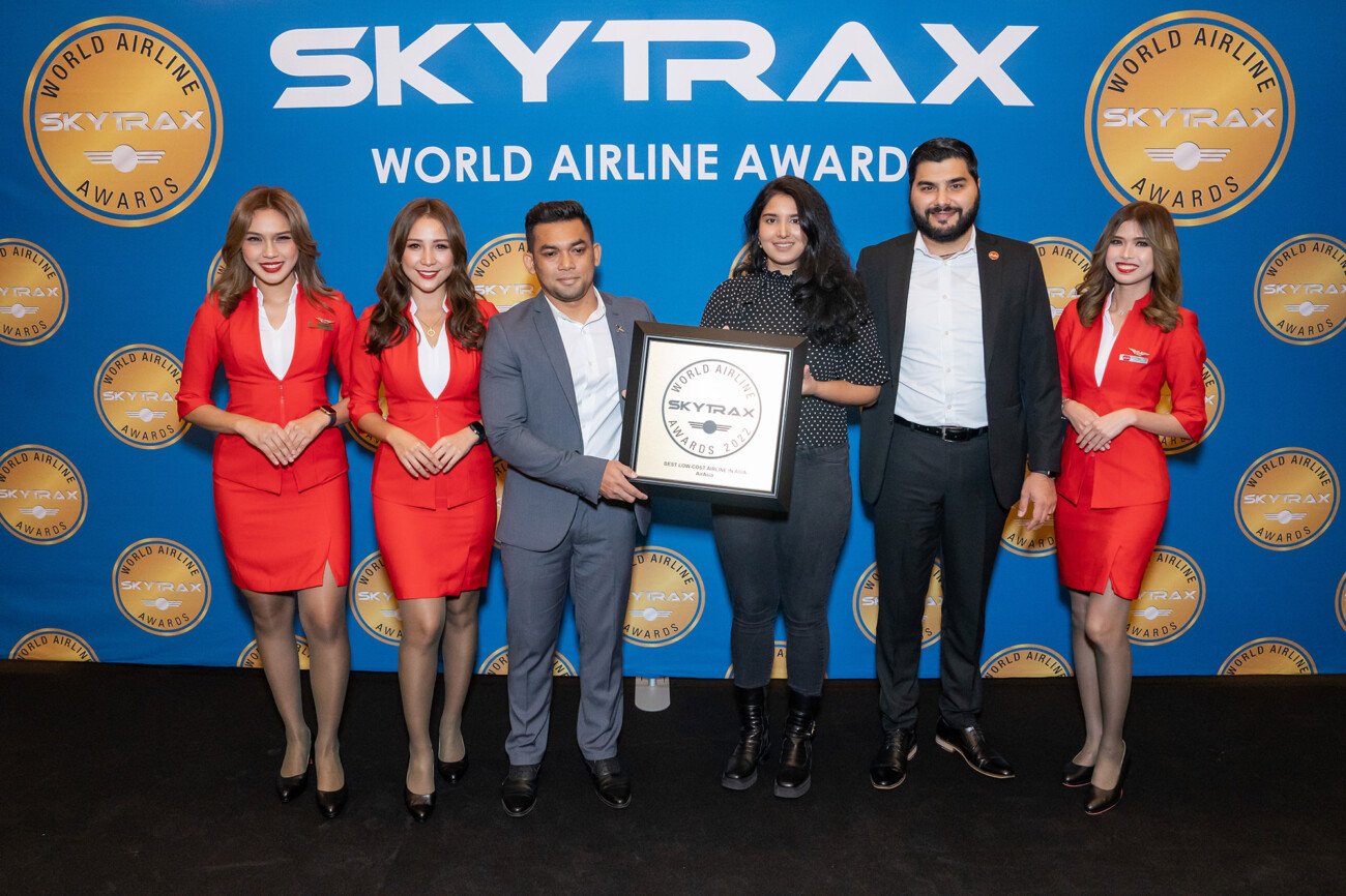 A group of six people is standing in front of a Skytrax World Airline Awards backdrop. Four individuals in red uniforms stand on the sides, while the two in the center hold a Skytrax award, smiling for the photo
