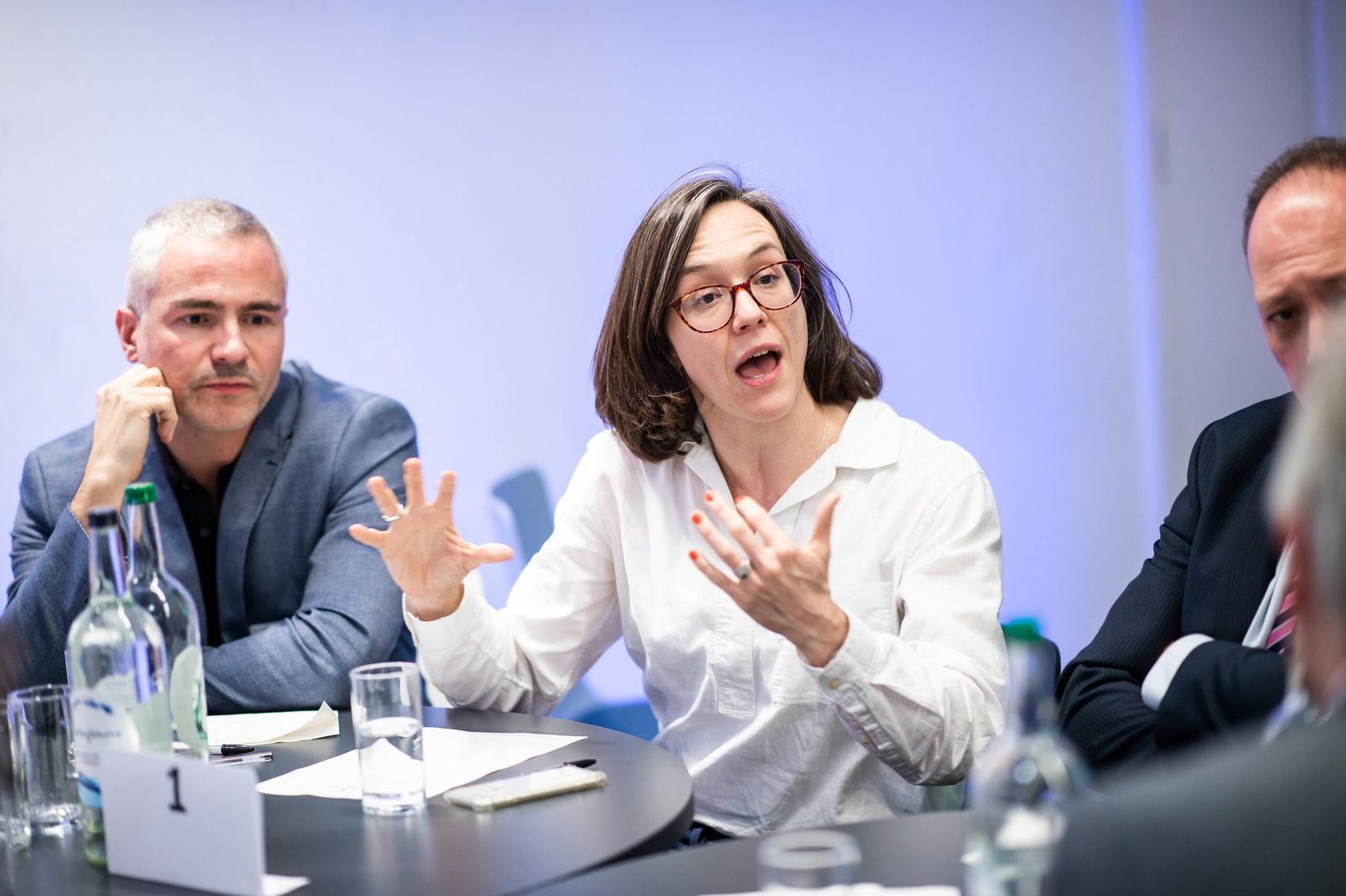 A woman with glasses and a white shirt speaks animatedly, gesturing with her hands during a discussion at a round table with two men and water bottles