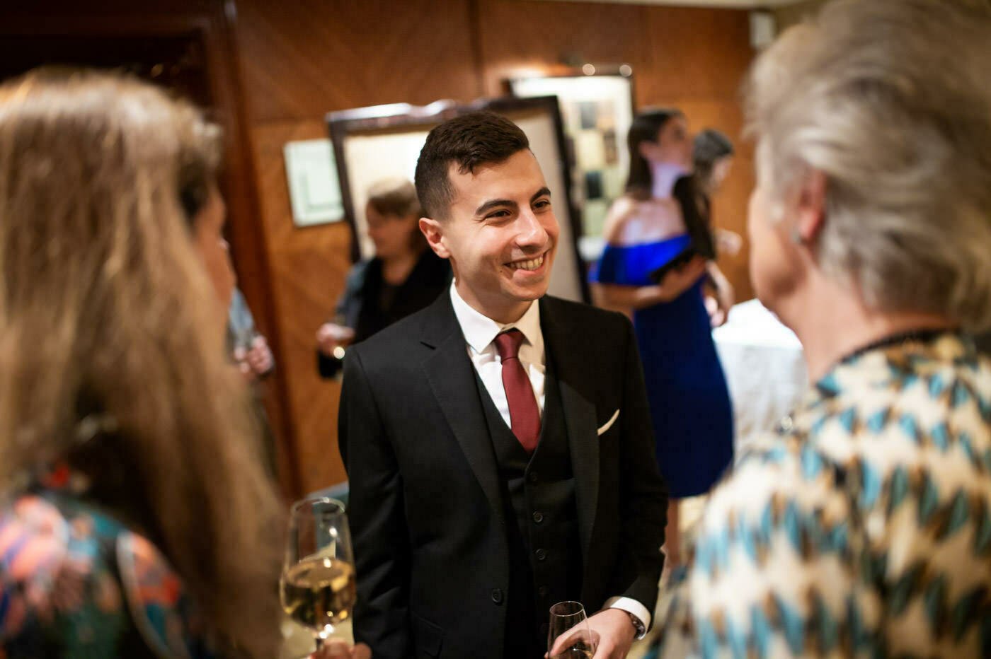 A young man in a suit and tie is smiling and holding a drink while talking to two women at a social event. Others are visible in the background, with soft, warm lighting creating an inviting atmosphere. James Gifford-Mead Photography - Event Photographer London