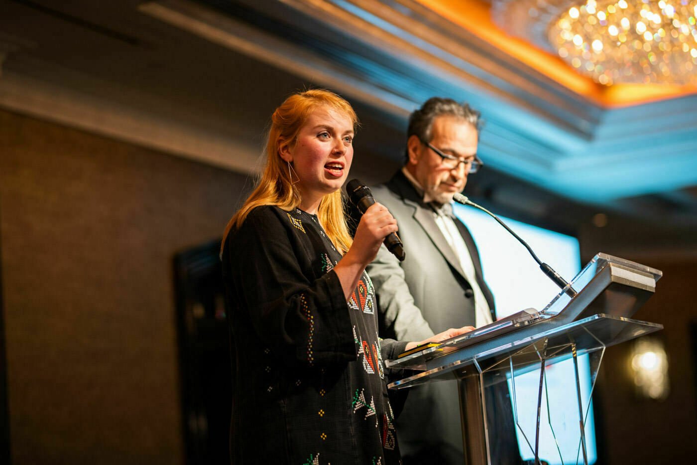 A woman with long hair speaks into a microphone at a podium, while a man in a suit and tie stands beside her reading from papers. They are under a chandelier in a warmly lit room, possibly during an event or presentation. James Gifford-Mead Photography - Event Photographer London