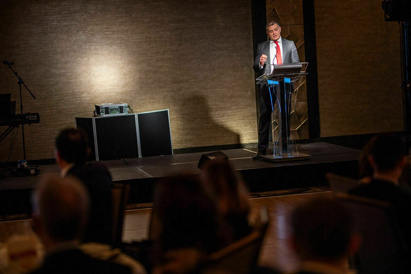 A person in a suit and red tie speaks at a clear podium on a stage, under focused lighting. An audience sits in the foreground, blurred. The background features textured walls and stage equipment. James Gifford-Mead Photography - Event Photographer London