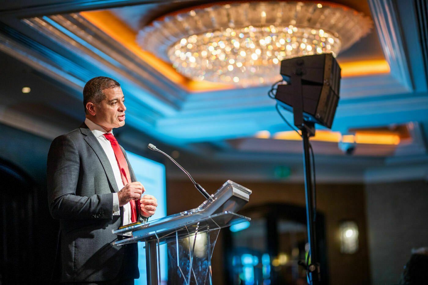 A person in a suit and red tie speaks at a podium with a microphone in a well-lit, elegant room. A chandelier and a screen are visible in the background