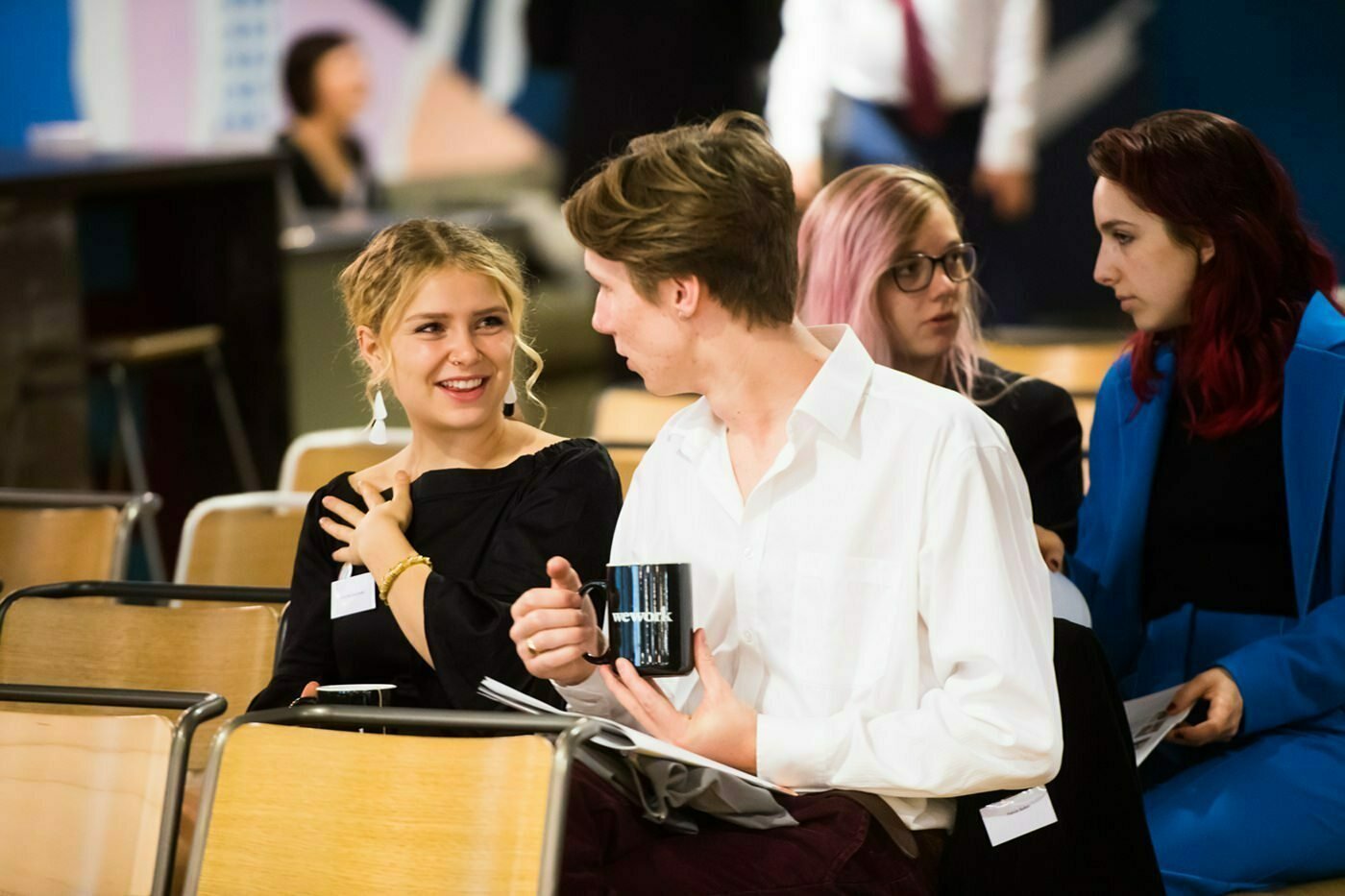 Four people sit in a room with rows of wooden chairs. A man and a woman in the foreground are talking, with the man holding a mug. Two women in the background are engaged in conversation. The setting appears to be informal or a casual event. James Gifford-Mead Photography - Event Photographer London