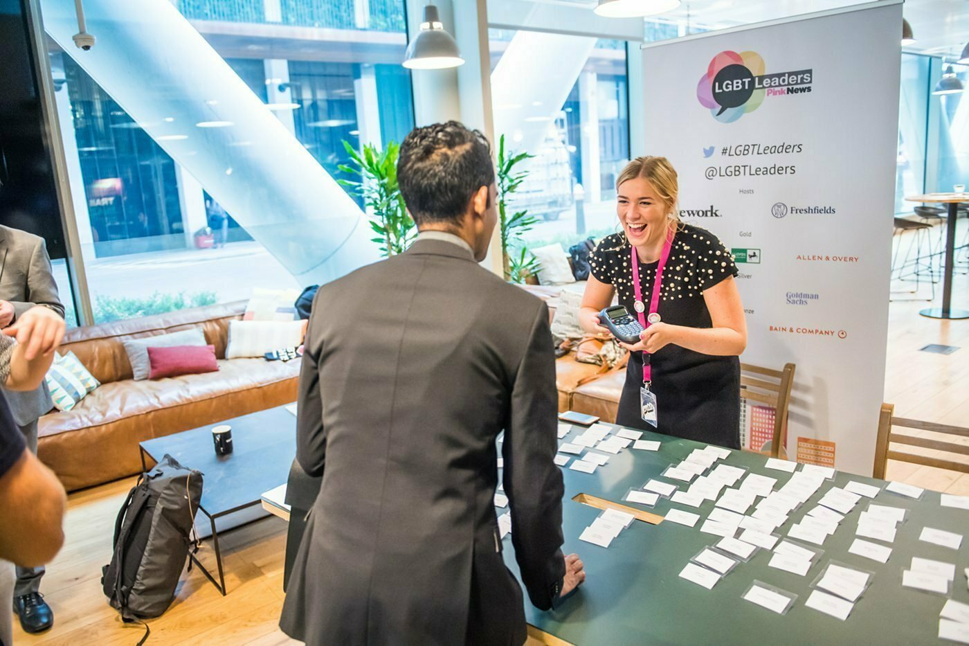 A person stands at a table covered with name badges, smiling at another person in a suit. A banner with "LGBT Leaders" and social media handles is visible. The setting appears to be a modern, open office space with large windows and a sofa. James Gifford-Mead Photography - Event Photographer London