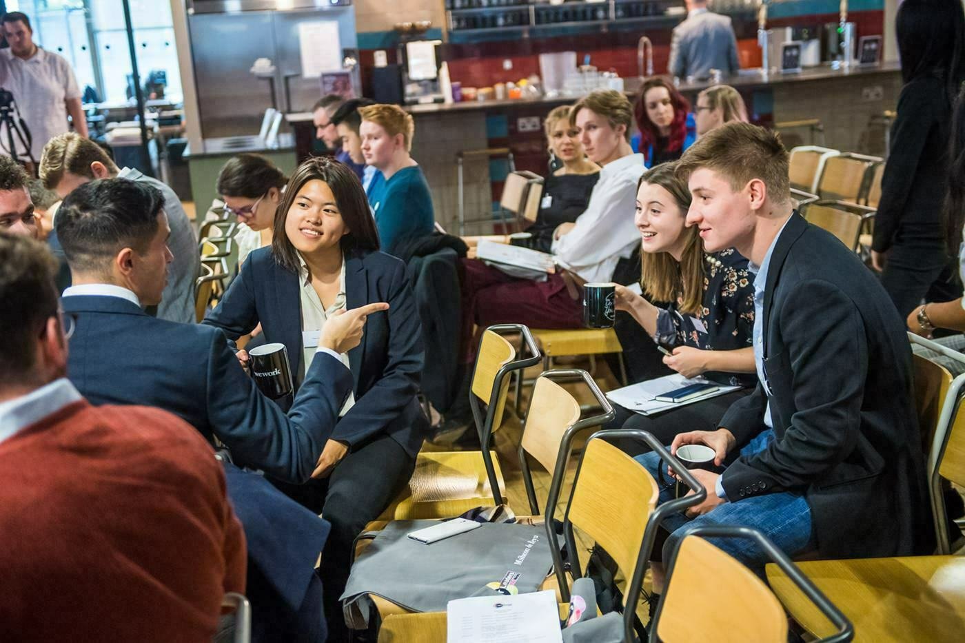 People are sitting in a meeting room, engaged in conversation. Some are holding coffee mugs and papers. The atmosphere is informal, with people smiling and interacting in small groups. The room has a mix of wooden chairs and modern decor. James Gifford-Mead Photography - Event Photographer London