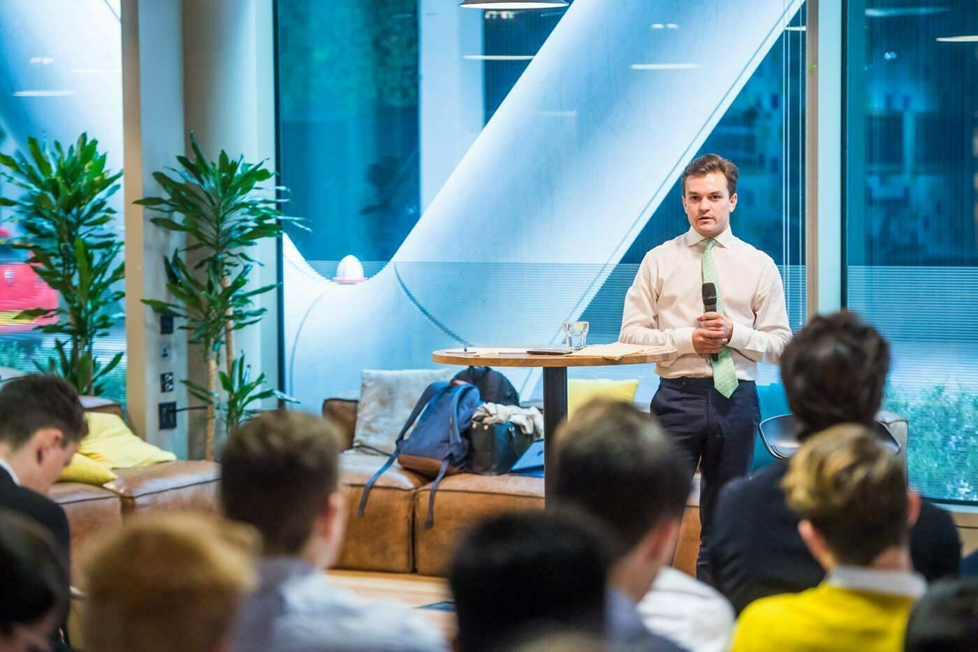 A man in a white shirt and tie stands holding a microphone, addressing a seated audience in a modern, brightly lit room. Large windows and green plants are in the background. James Gifford-Mead Photography - Event Photographer London