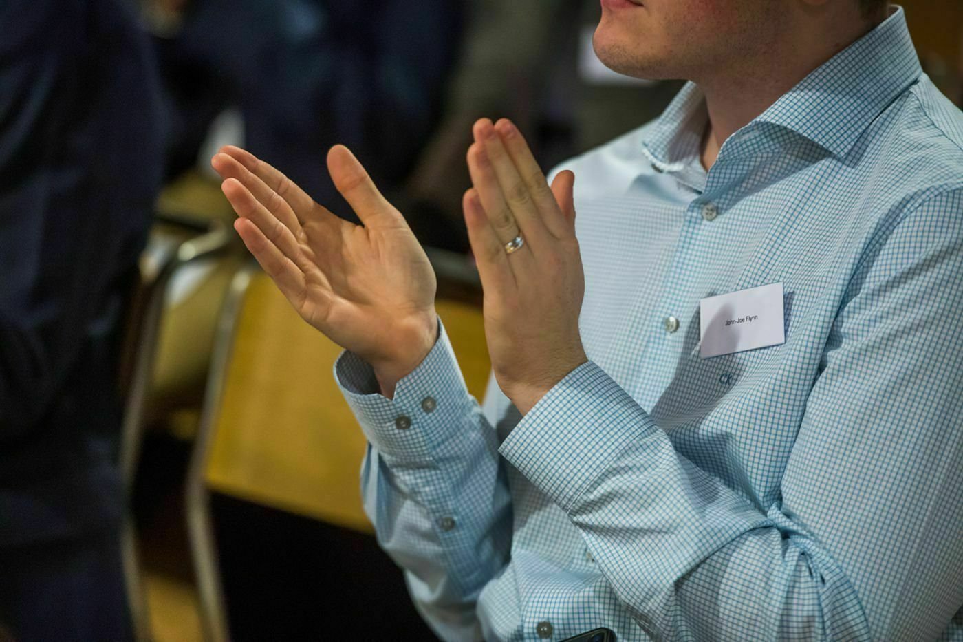 A person in a light blue checkered shirt is sitting and clapping. They have a nametag on their chest, though the name is unclear. The background shows partially visible chairs and another person wearing a dark outfit. James Gifford-Mead Photography - Event Photographer London