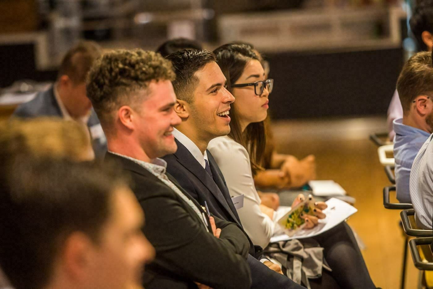 People seated in a row, facing forward, and attentively watching something. They are dressed in business attire, with some holding papers. They appear to be engaged and smiling, suggesting a conference or meeting setting. James Gifford-Mead Photography - Event Photographer London
