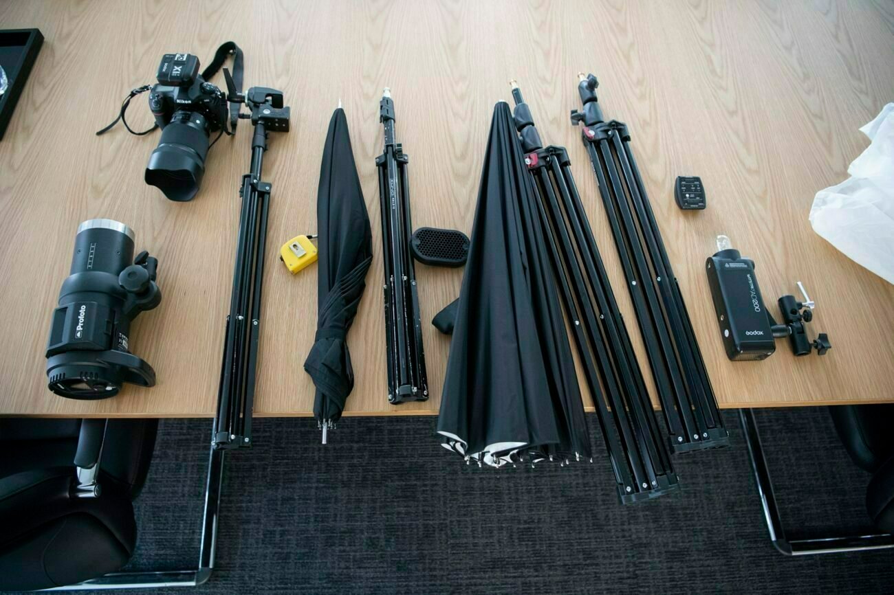 Photography equipment spread on a wooden table, including a camera, lenses, tripods, light stands, umbrellas, a tape measure, and a remote trigger. The items are neatly arranged in rows, suggesting preparation for a photoshoot. James Gifford-Mead Photography - Event Photographer London