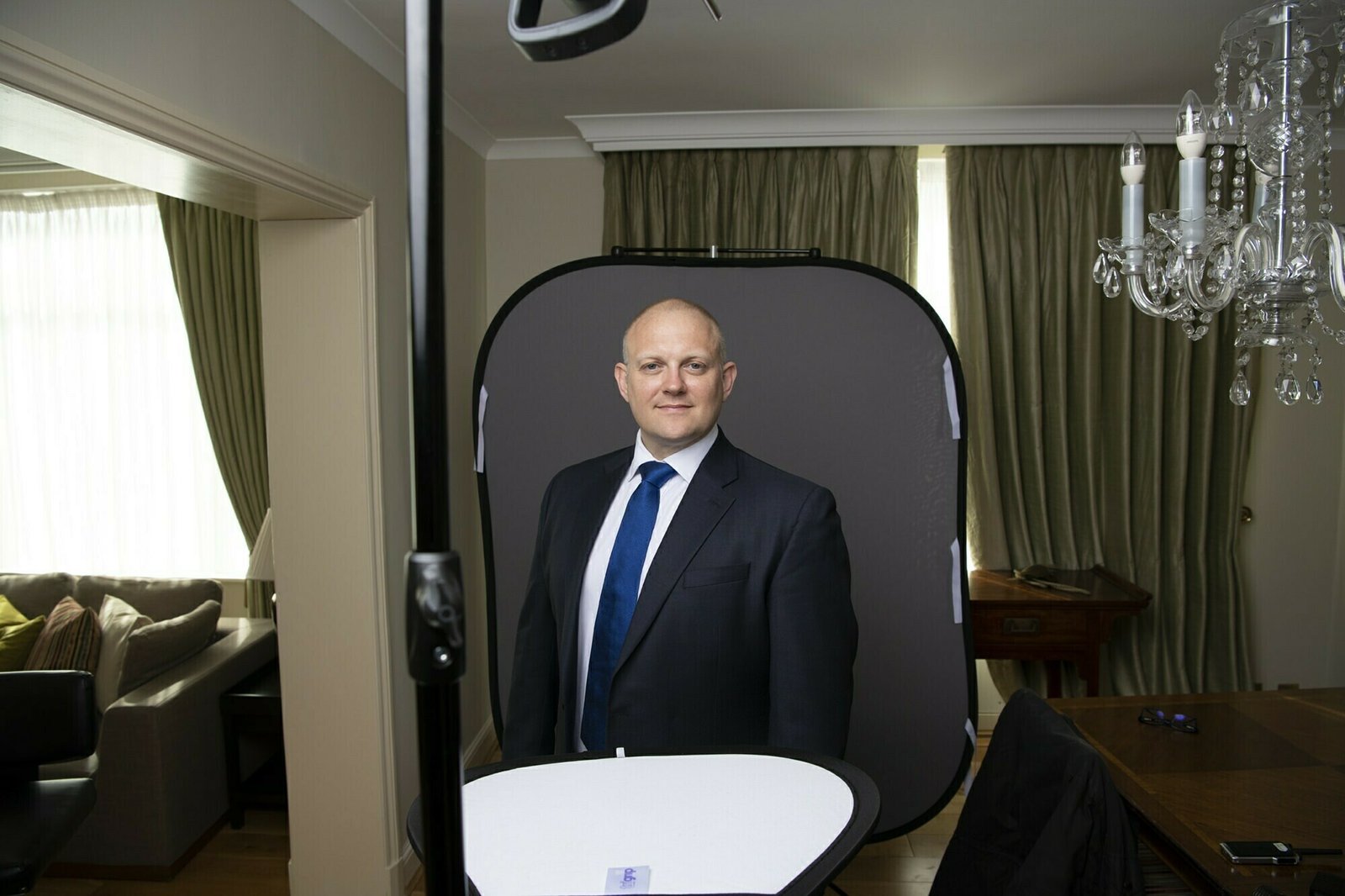 A man in a suit and blue tie stands in front of a studio backdrop, set up in a well-lit room with curtains and a chandelier. Photography equipment is visible in the foreground. James Gifford-Mead Photography - Event Photographer London