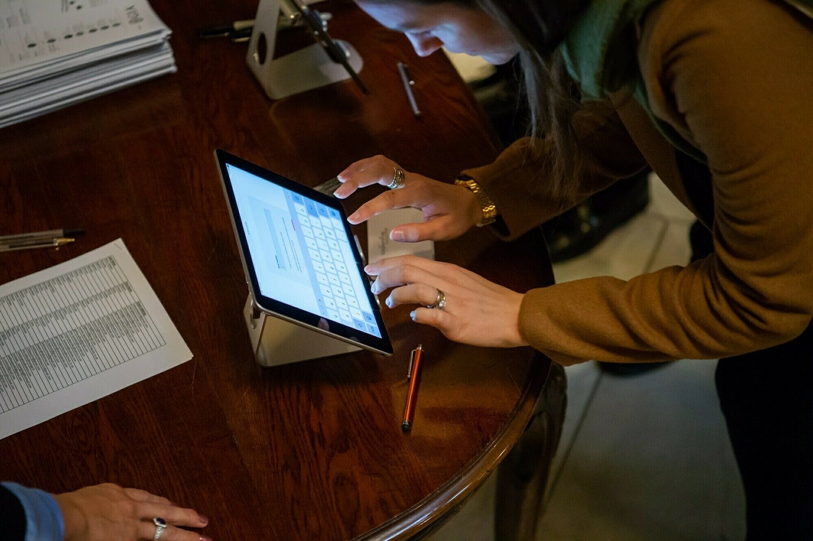 A person in a brown jacket is using a tablet placed on a wooden table, typing on the screen. Papers, a pen, and metallic stands are nearby. The scene appears to be a professional or office setting. James Gifford-Mead Photography - Event Photographer London