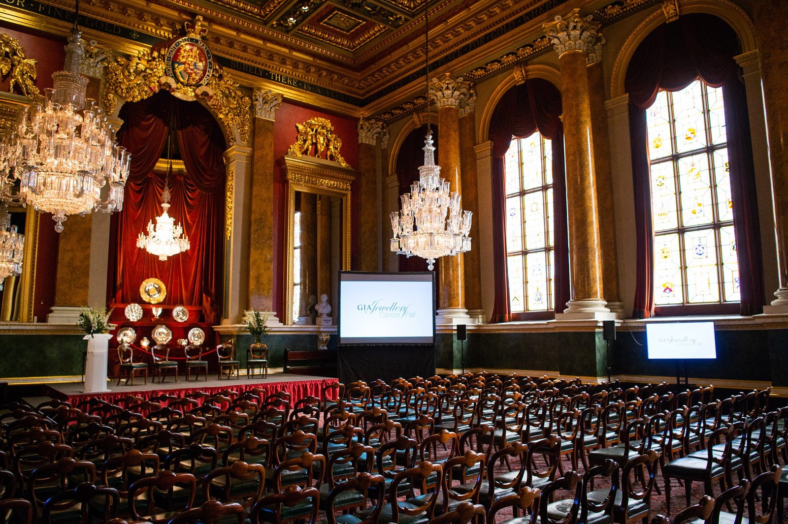 An ornate hall with chandeliers and stained glass windows. Rows of chairs face a stage with red drapery and a podium. Two large screens display "30 Year Anniversary" on each side of the stage. James Gifford-Mead Photography - Event Photographer London