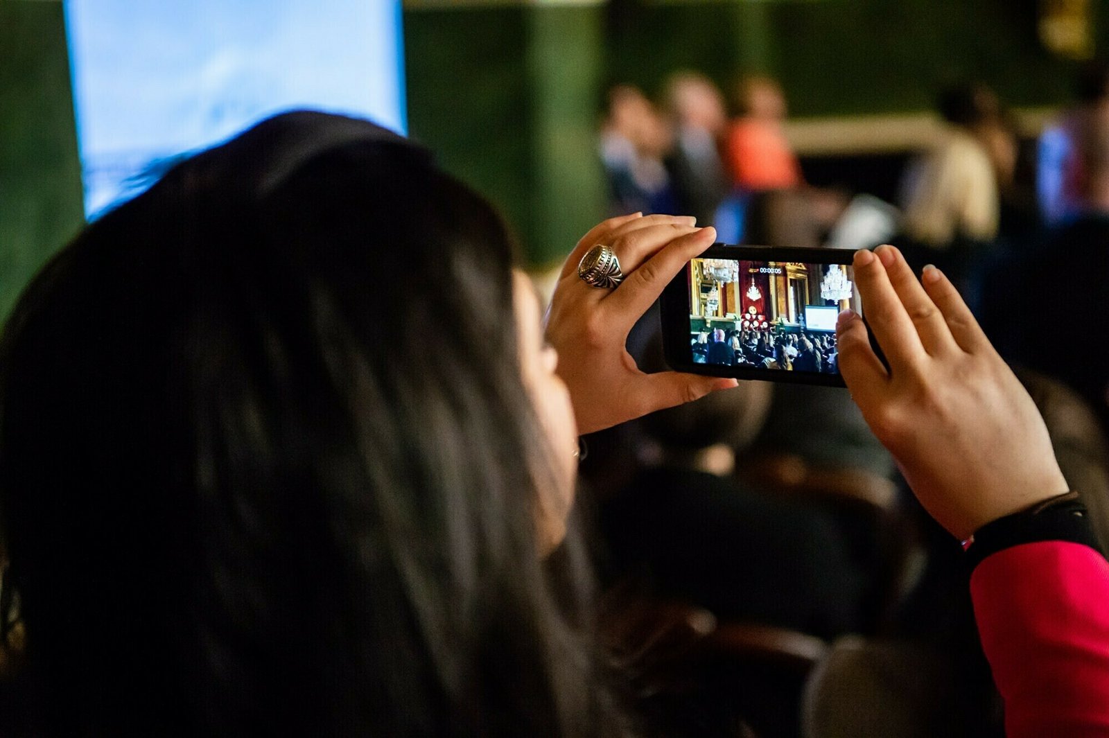 A person with long dark hair is holding a smartphone, capturing a photo or video of a formal event or gathering. The background is blurred, showing several people seated and some standing near a stage or presentation area. James Gifford-Mead Photography - Event Photographer London