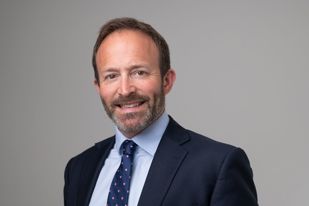 A man with short dark hair and a beard is smiling while wearing a dark suit, white shirt, and polka dot tie. The background is plain light gray. James Gifford-Mead Photography - Event Photographer London