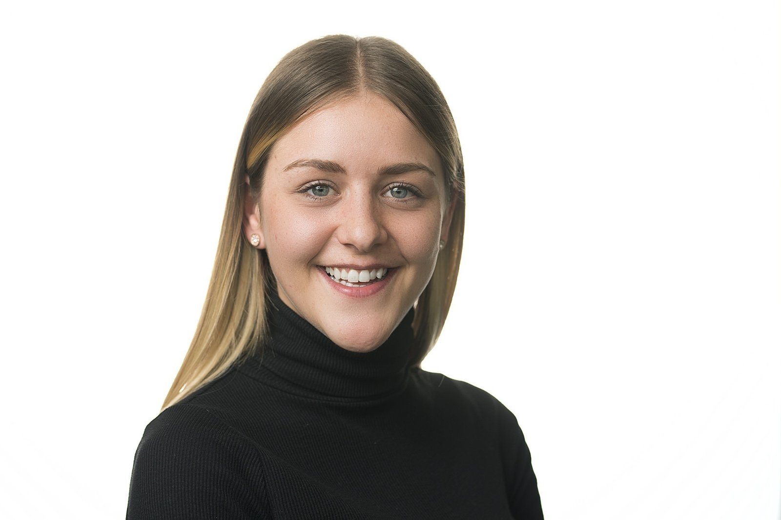A woman with long brown hair, wearing a black turtleneck and pearl earrings, smiles at the camera against a plain white background