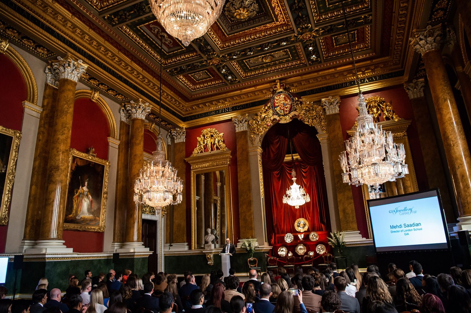 A large, ornate room with high ceilings, crystal chandeliers, and gold-accented decor is filled with people seated in rows. A presentation is being shown on a screen, with a speaker standing at a podium. Rich curtains and framed paintings adorn the walls. James Gifford-Mead Photography - Event Photographer London