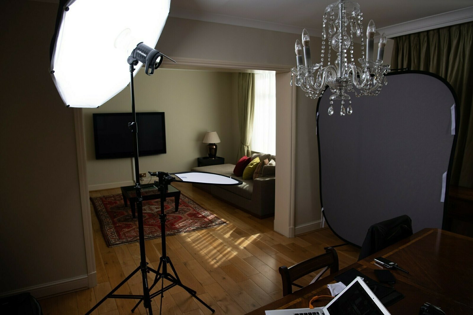 A living room setup for a photo shoot with a large softbox light, a reflector, and a gray backdrop. There is a chandelier hanging from the ceiling, and a TV mounted on the wall. A laptop and papers are visible on the dining table. James Gifford-Mead Photography - Event Photographer London