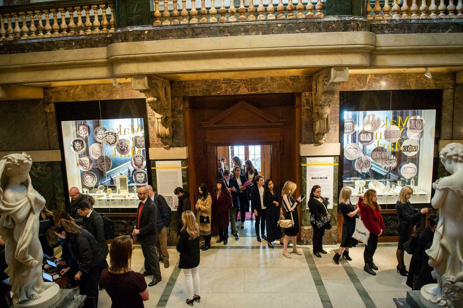 A group of people walk and gather around in a museum with ornate decor. Displays with various objects and information are visible on the walls. Two large sculptures are in the foreground. The atmosphere is busy and lively. James Gifford-Mead Photography - Event Photographer London