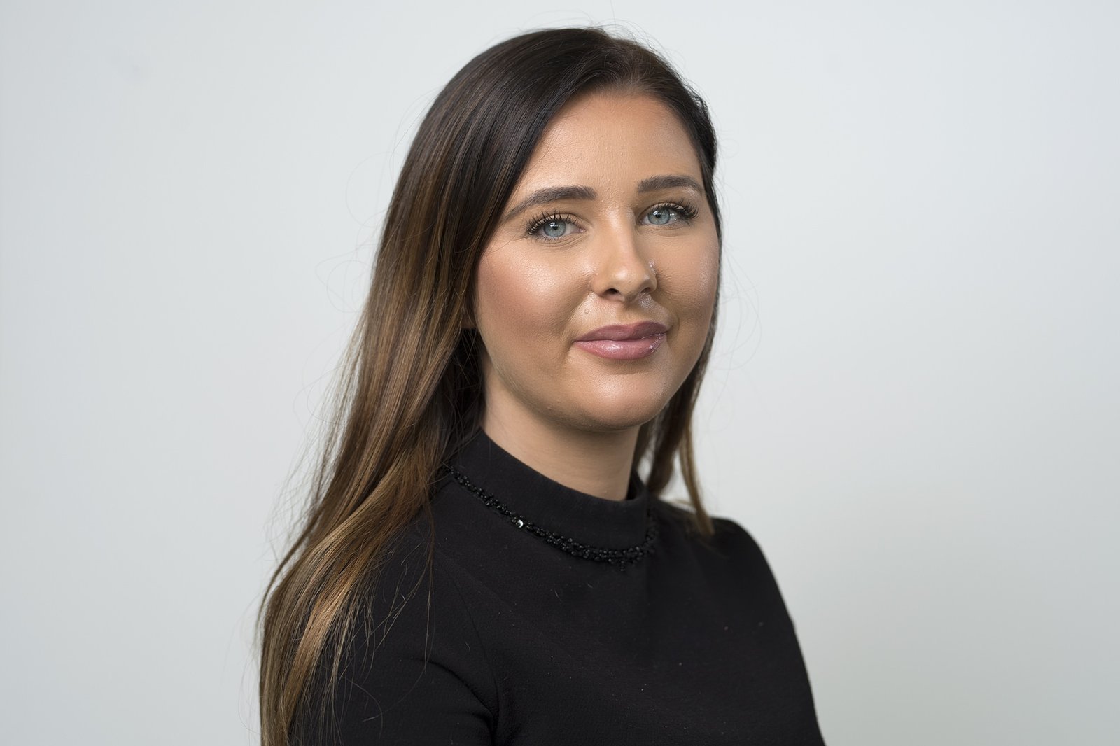 A woman with long brown hair and blue eyes is smiling slightly. She is wearing a black top and is posed against a plain white background. James Gifford-Mead Photography - Event Photographer London