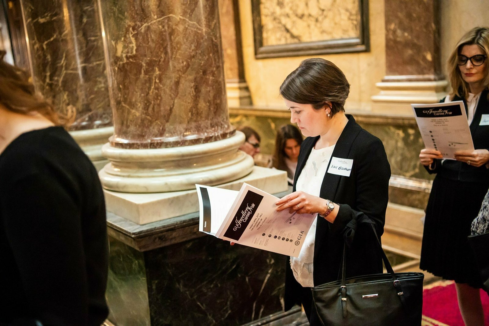 A group of people stand indoors near marble columns, focusing on brochures. A woman in the foreground, wearing a black blazer and a name tag, looks intently at her brochure. The setting appears formal and ornate. James Gifford-Mead Photography - Event Photographer London