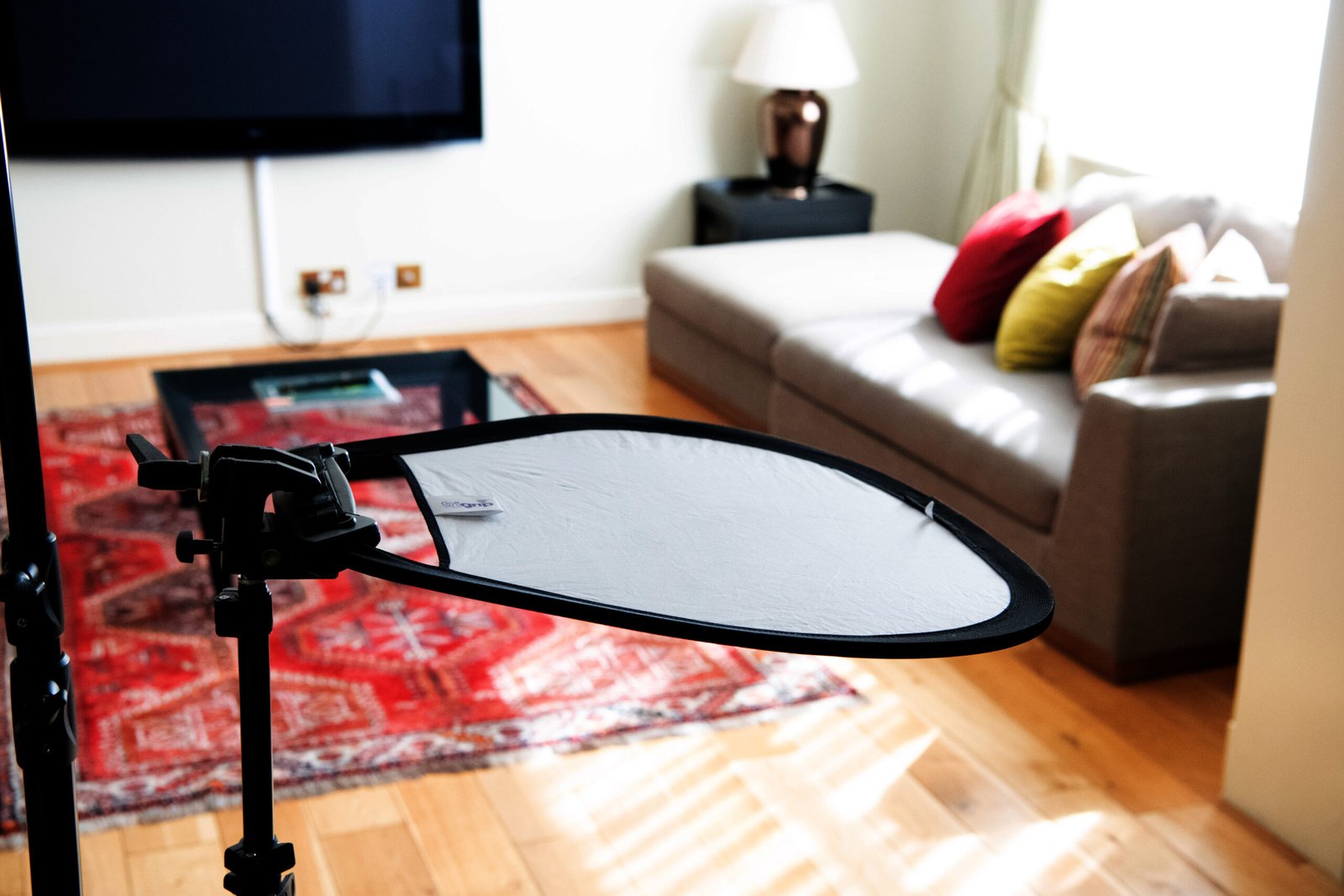 A photography reflector is set up in a bright living room with a light wood floor. A gray sofa with colorful cushions is placed beside a large window. A red patterned rug lies near a glass coffee table, and a flat-screen TV is mounted on the wall. James Gifford-Mead Photography - Event Photographer London