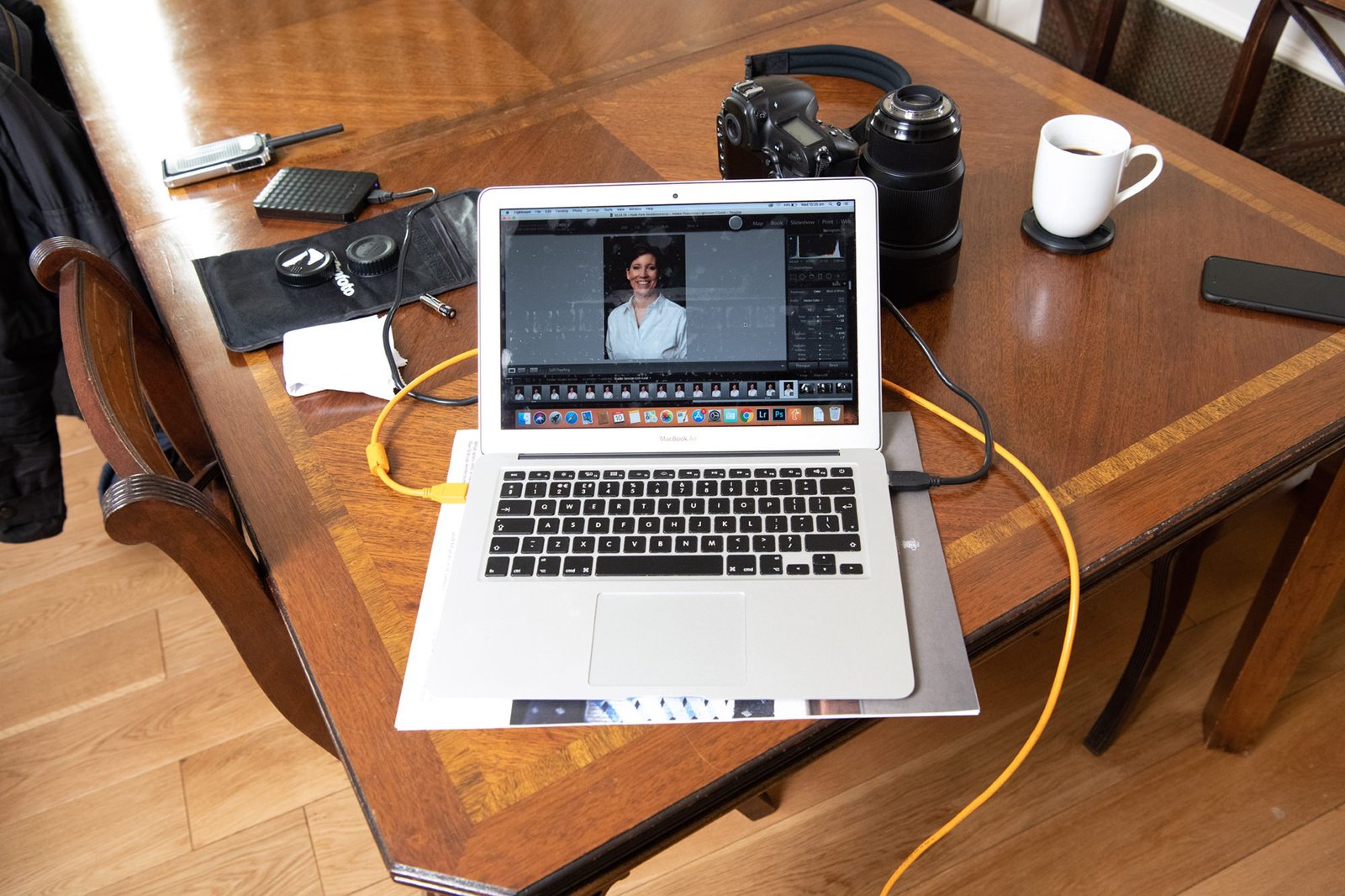 A laptop displaying a portrait photograph on its screen is on a wooden table. Nearby are a camera, camera lens, notepad, pen, and a cup of coffee. A yellow cable connects the laptop to a device off-screen. James Gifford-Mead Photography - Event Photographer London