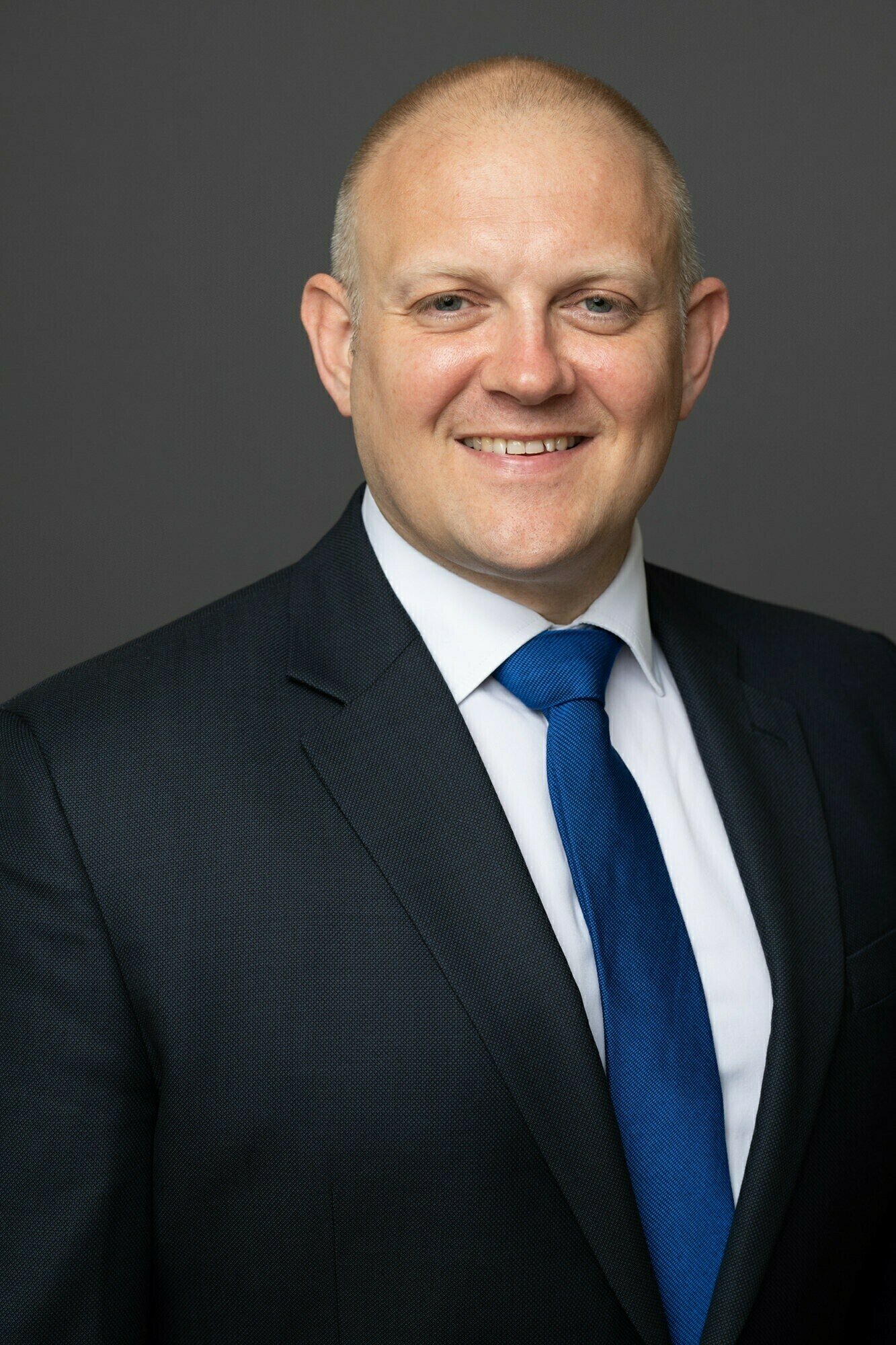 A bald man in a dark suit with a white shirt and blue tie is smiling. He stands against a plain gray background. James Gifford-Mead Photography - Event Photographer London