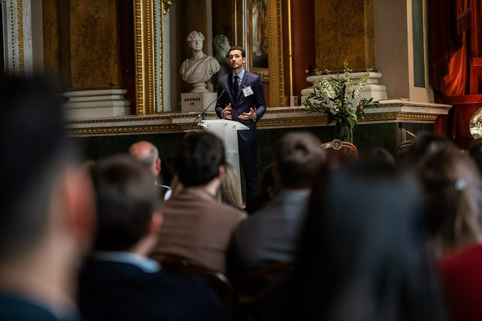 A person in a suit speaks at a podium in a grand room with ornate decor, including columns and a bust. An audience listens attentively, and there is a large floral arrangement visible to the side. James Gifford-Mead Photography - Event Photographer London