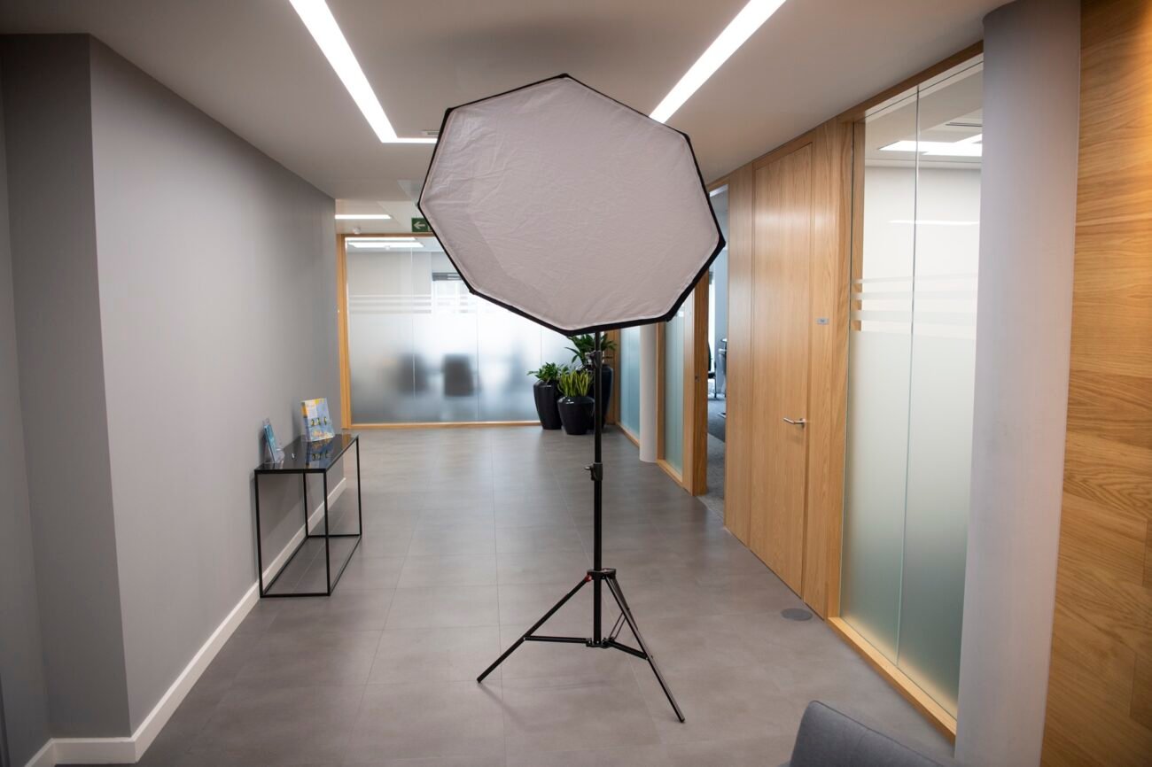 A photography softbox light is set up in a modern office hallway with gray walls and a tiled floor. The corridor is lined with wooden doors and glass panels, and a small table holds brochures near potted plants. James Gifford-Mead Photography - Event Photographer London