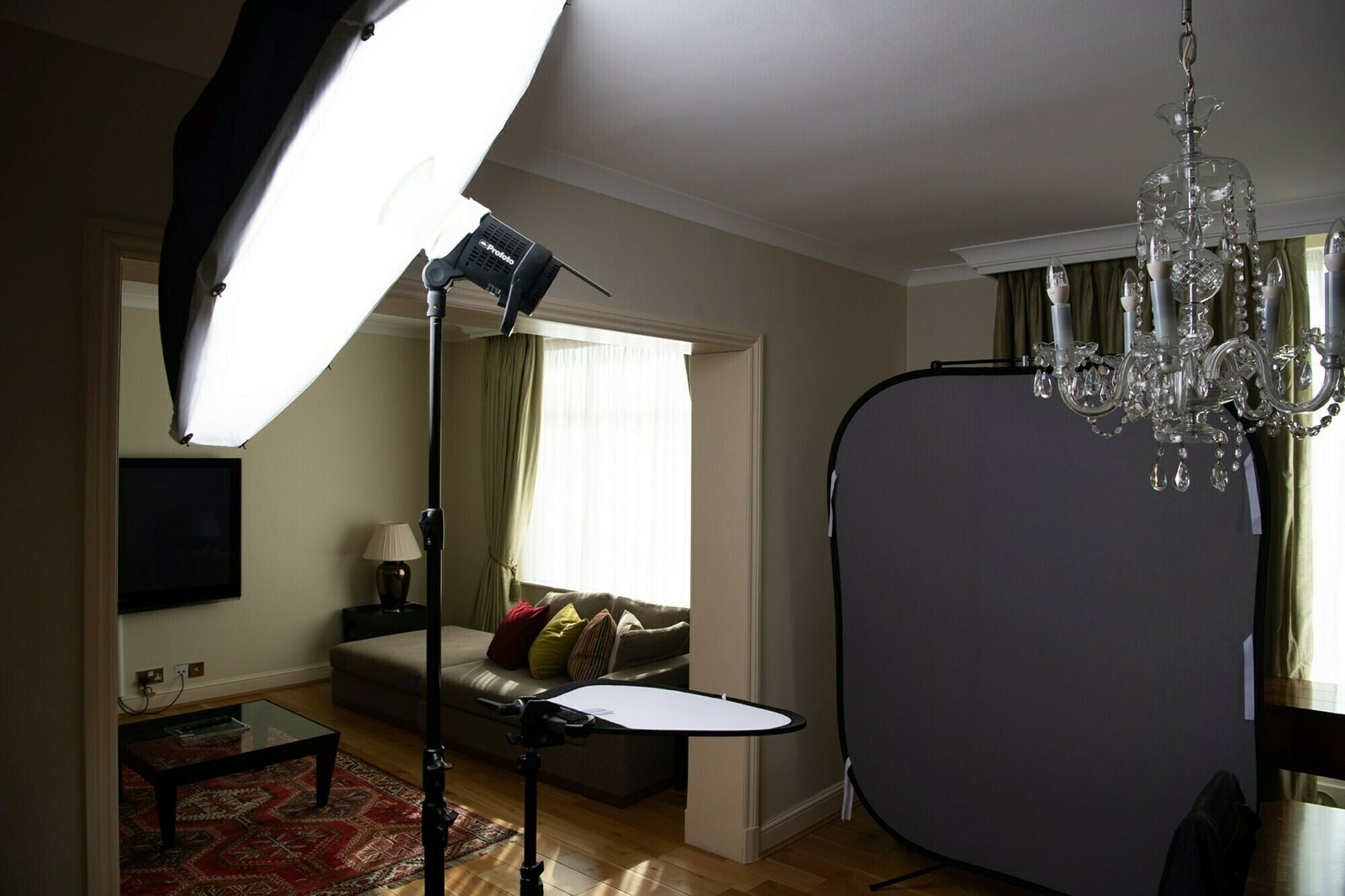 A home living room set up as a photo studio with a large softbox light, a reflector, and a backdrop. A chandelier is hanging from the ceiling, and a couch with colorful cushions is in the background. James Gifford-Mead Photography - Event Photographer London