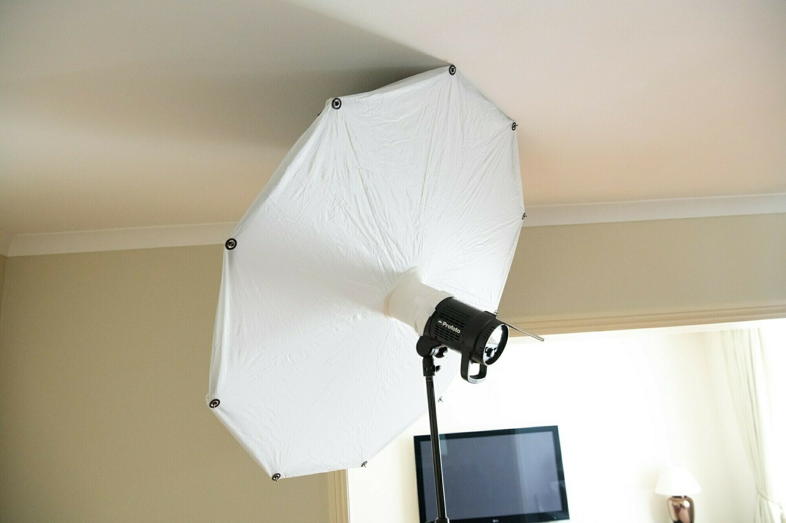 A large white photography umbrella light is set up in a room with beige walls and a white ceiling. A black TV is mounted on the wall in the background, and a lamp is visible to the side. James Gifford-Mead Photography - Event Photographer London