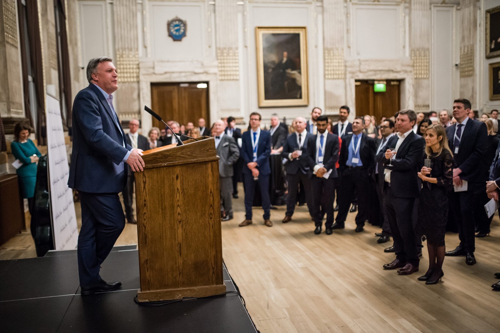 A man in a suit speaks at a wooden podium to a crowd in a formal room with ornate details and paintings. The audience, wearing business attire, listens attentively while standing
